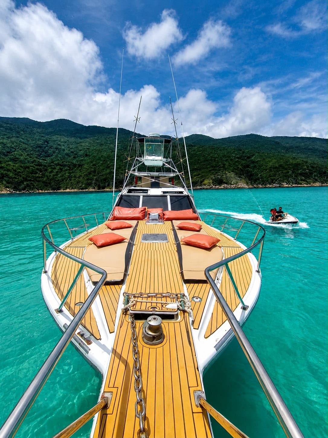 Proa da lancha com solário e almofadas laranja em águas turquesa de Arraial do Cabo, com um jet ski ao fundo